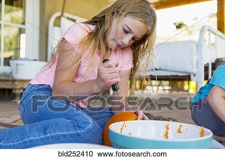 Stock Image - Caucasian girl sitting on floor carving pumpkin with knife. Fotosearch