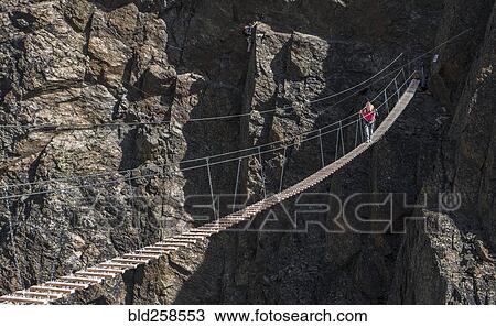 Stock Image - Caucasian man crossing rope bridge on mountain. Fotosearch