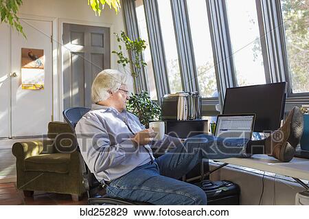 Stock Photo - Caucasian man drinking coffee and using laptop with feet up on desk. Fotosearch