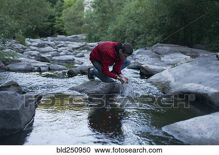 Stock Image - Caucasian man kneeling on rock in river cupping water with hands. Fotosearch