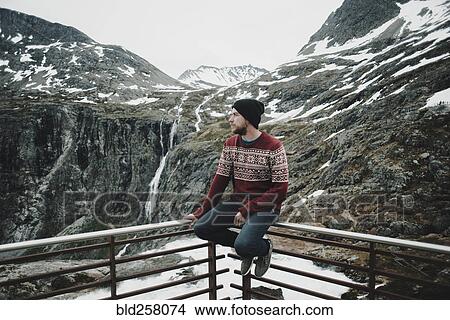 Picture - Caucasian man sitting on balcony railing admiring snow on mountain. Fotosearch