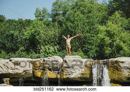 Stock Image - Caucasian man standing on rocks near waterfalls. Fotosearch