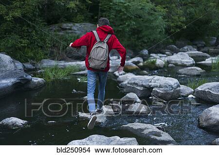 Picture - Caucasian man walking on rocks crossing river. Fotosearch