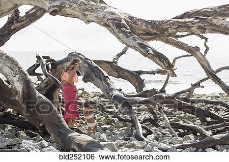 Stock Photograph - Caucasian mother and daughter climbing on driftwood at beach. Fotosearch