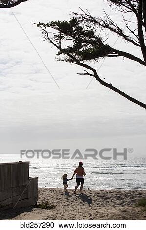 Stock Image - Caucasian mother and daughter holding hands and running on beach. Fotosearch
