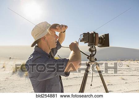 Stock Image - Caucasian photographer with tripod shielding eyes in desert. Fotosearch