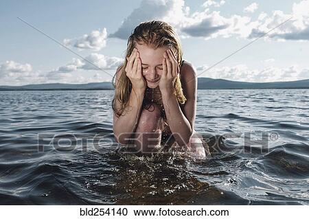 Stock Image - Caucasian teenage girl kneeling in ocean splashing water on face. Fotosearch