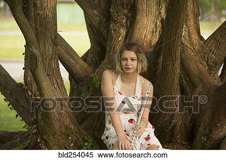 Stock Photography - Caucasian teenage girl sitting on tree root holding flute. Fotosearch
