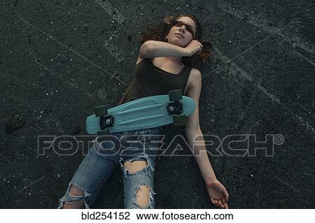 Stock Image - Caucasian teenage girl with skateboard laying on pavement covering eyes. Fotosearch