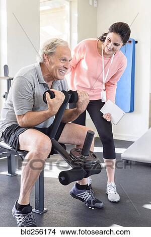 Picture - Caucasian trainer watching man strengthen arms. Fotosearch