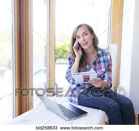 Stock Image - Caucasian woman holding paperwork and talking on cell phone near window. Fotosearch