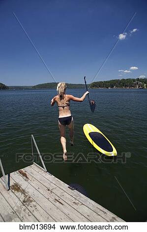Picture - Caucasian woman jumping in lake next to paddleboard. Fotosearch