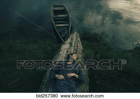 Stock Image - Caucasian woman laying on upside-down boat near lake. Fotosearch