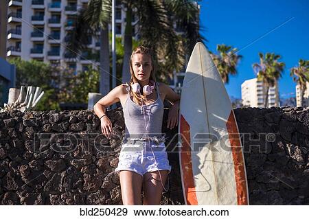 Stock Photo - Caucasian woman leaning on stone wall near surfboard. Fotosearch