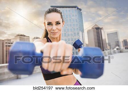Stock Photo - Caucasian woman lifting dumbbells on urban rooftop. Fotosearch