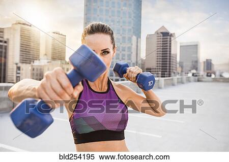 Stock Photography - Caucasian woman lifting dumbbells on urban rooftop. Fotosearch