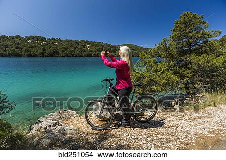 Picture - Caucasian woman on bicycle photographing lake with cell phone. Fotosearch