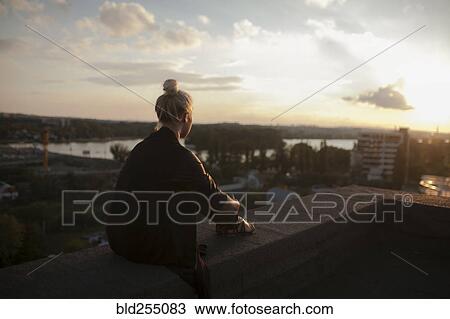 Stock Image - Caucasian woman on roof admiring scenic view of sunset. Fotosearch