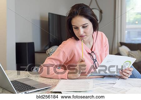 Picture - Caucasian woman reading paperwork near laptop. Fotosearch