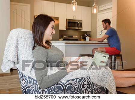 Stock Image - Caucasian woman sitting in armchair using laptop and drinking coffee. Fotosearch
