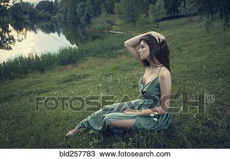 Stock Image - Caucasian woman sitting in grass with wildflowers near lake. Fotosearch
