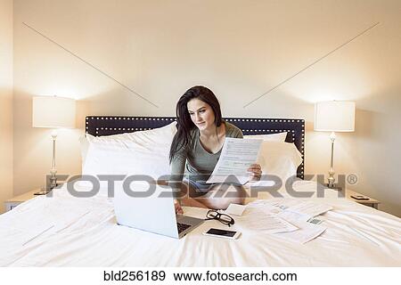 Caucasian woman sitting on bed using laptop near paperwork View Large Photo Image Stock Photo - Caucasian woman sitting on bed using laptop near paperwork. Fotosearch