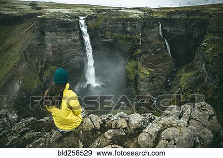 Stock Photo - Caucasian woman sitting on cliff admiring waterfalls. Fotosearch
