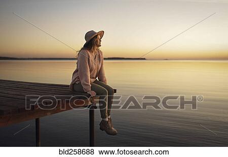 Stock Photo - Caucasian woman sitting on dock of lake admiring sunset. Fotosearch