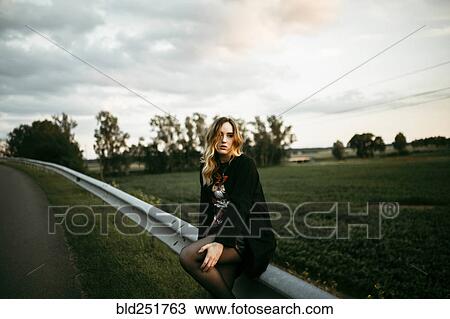 Stock Image - Caucasian woman sitting on guardrail near road. Fotosearch