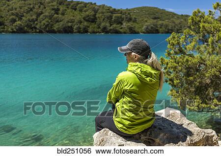 Stock Photograph - Caucasian woman sitting on rock at lake. Fotosearch