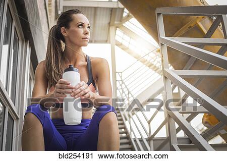 Stock Image - Caucasian woman sitting on urban staircase holding water bottle. Fotosearch
