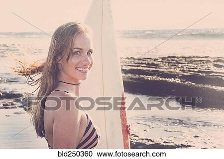 Stock Image - Caucasian woman standing on beach holding surfboard. Fotosearch