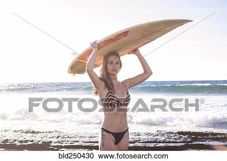 Stock Image - Caucasian woman standing on beach holding surfboard. Fotosearch