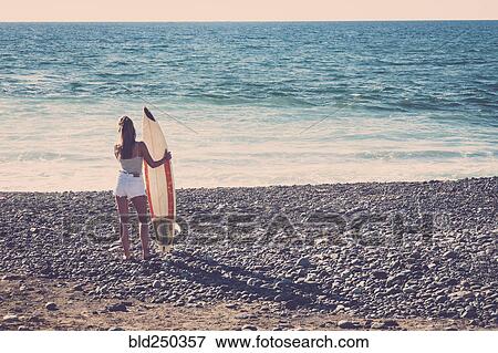Stock Photo - Caucasian woman standing on beach holding surfboard. Fotosearch