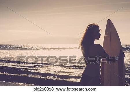 Stock Photo - Caucasian woman standing on beach holding surfboard. Fotosearch
