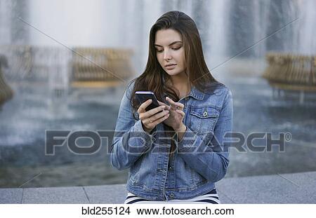 Picture - Caucasian woman texting on cell phone at fountain. Fotosearch