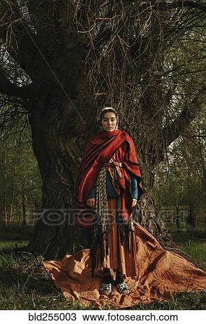 Stock Image - Caucasian woman wearing traditional clothing standing near tree. Fotosearch