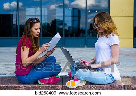 Stock Photo - Caucasian women reading paperwork and using laptop outdoors. Fotosearch