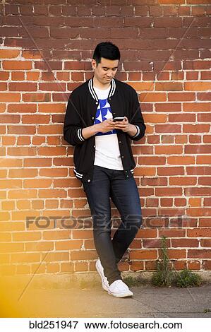 Stock Photo - Chinese man leaning on brick wall texting on cell phone. Fotosearch