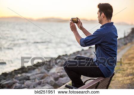 Stock Image - Chinese man sitting on bench photographing ocean with cell phone. Fotosearch