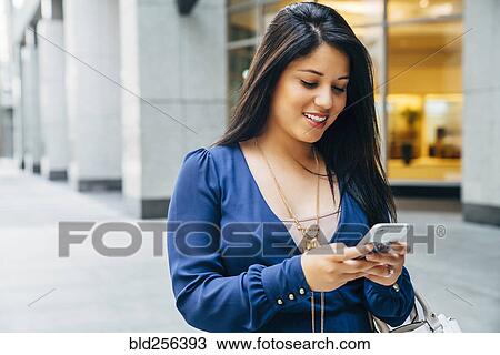 Stock Image - Close up of Hispanic woman texting on cell phone in city. Fotosearch