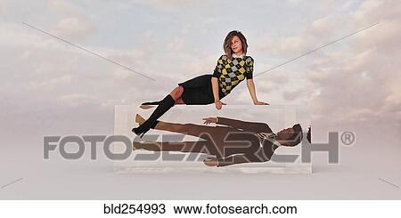 Stock Image - Confident businesswoman sitting on cube with businessman trapped inside. Fotosearch
