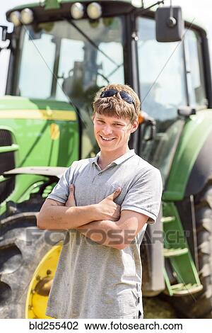 Stock Image - Confident Caucasian man standing near tractor. Fotosearch