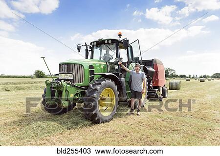 Stock Image - Confident Caucasian man standing near tractor. Fotosearch