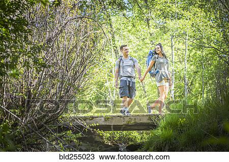 Stock Image - Couple carrying backpacks across bridge in woods. Fotosearch