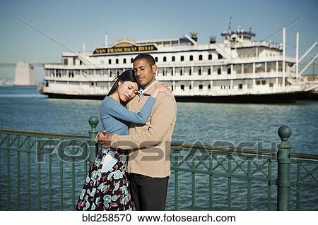Stock Image - Couple hugging at waterfront near boat. Fotosearch
