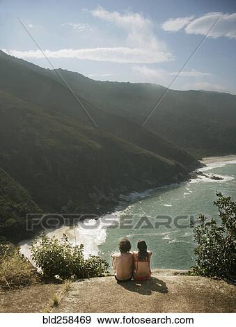 Couple sitting on cliff over ocean beach View Large Photo Image Stock Photo - Couple sitting on cliff over ocean beach. Fotosearch