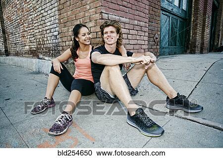 Stock Image - Couple sitting on sidewalk leaning on corner of brick building. Fotosearch
