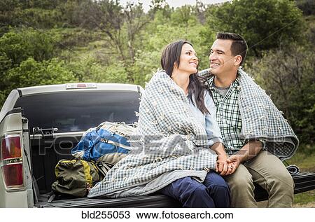 Stock Image - Couple wrapped in blanket sitting on bed of pick-up truck. Fotosearch