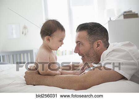 Stock Image - Father playing with baby son sitting on bed. Fotosearch